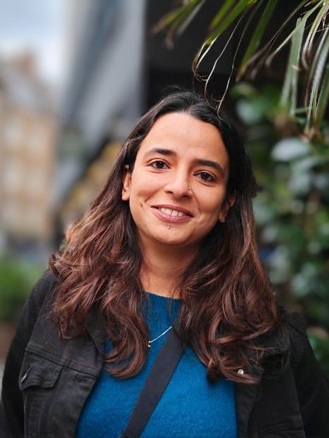 Photo of Akriti Mehta. A woman with long, wavy brown hair smiling at the camera. She is wearing a blue top, and a black jacket. The background is softly blurred, showing greenery and an urban setting with buildings and plants.