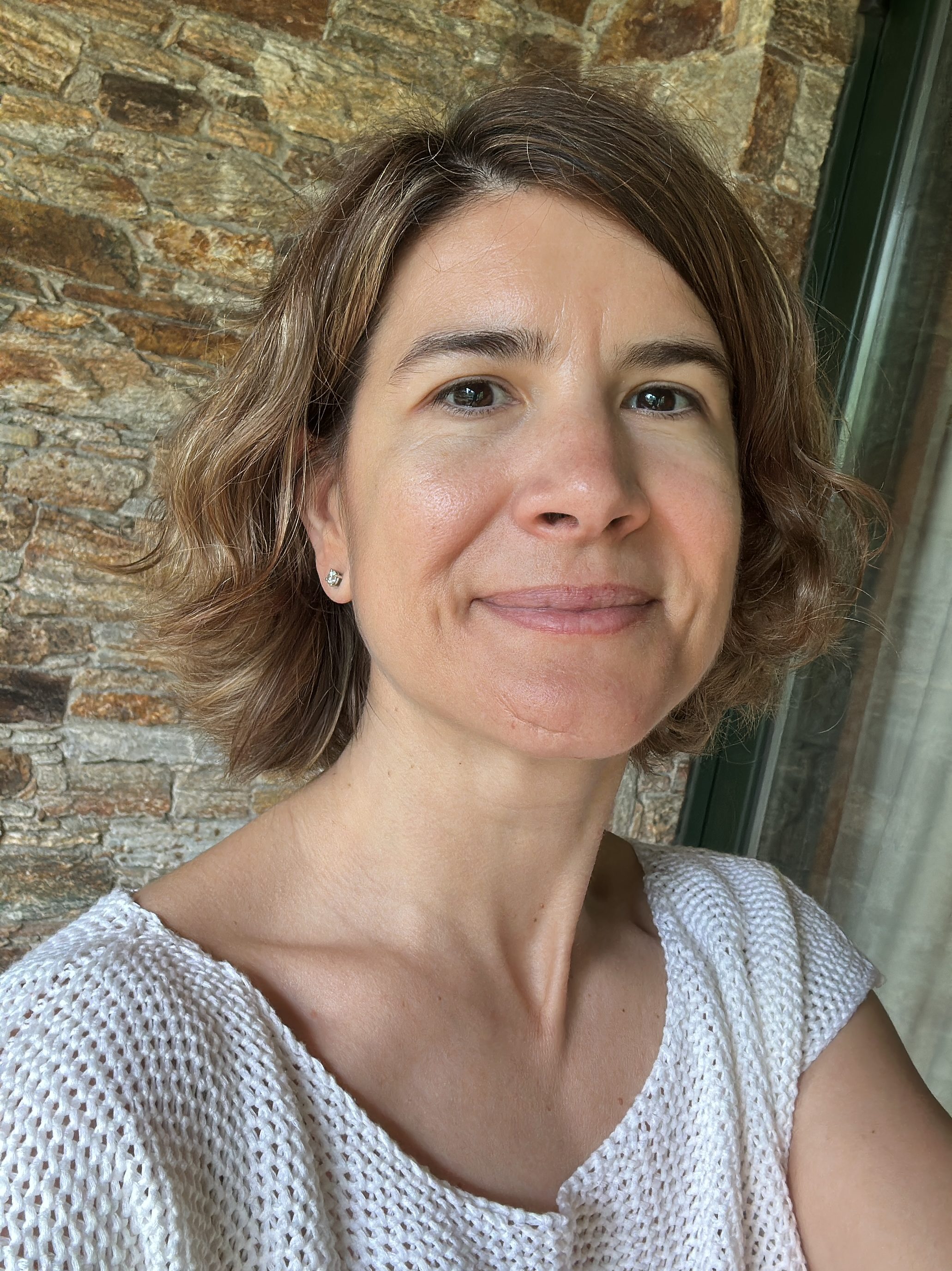 Photo of Mercedes Ramiro, a woman with short brown hair wearing a white knit top, smiling slightly in front of a stone wall.