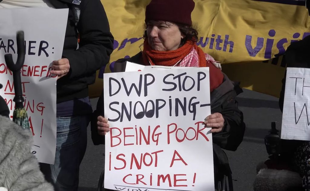 A woman in a wheelchair at a protest holds up a sign that says "DWP Stop Snooping - Being poor is not a crime". She is surrounded by other protestors.