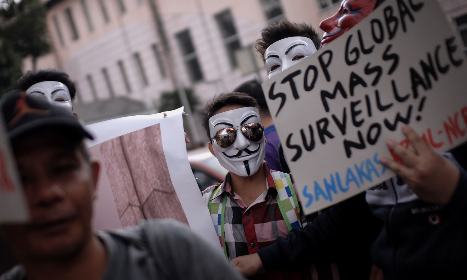A person in a Guy Fawkes mask at a protest looks into the camera. A second person holds up a sign saying "Stop Global Mass Surveillance Now".