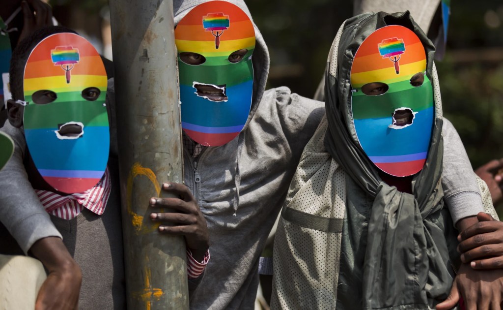 Three people are standing with their arms around each other's shoulders. They are wearing full face masks with only their eyes and lips visible. The masks are rainbow coloured.