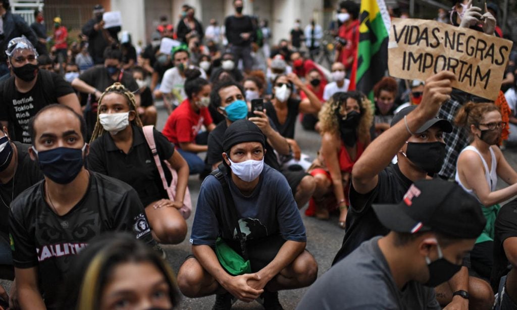 A group of people are kneeling. A person holds up a sign reading "Vidas Negras Importam".