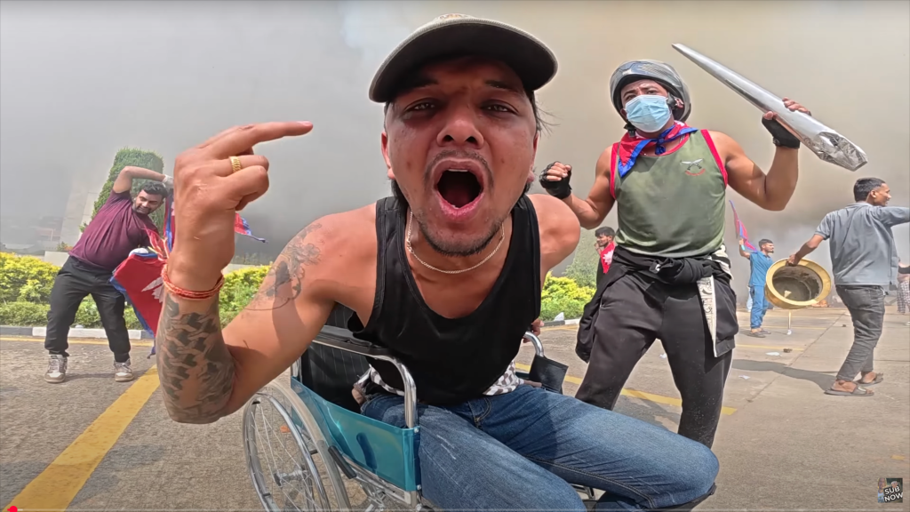 A young disabled man in a wheelchair leans into the camera. He is shouting, raising his middle finger, and his body language is defiant. In the background, several men can be seen carrying items amidst plumes of smoke.