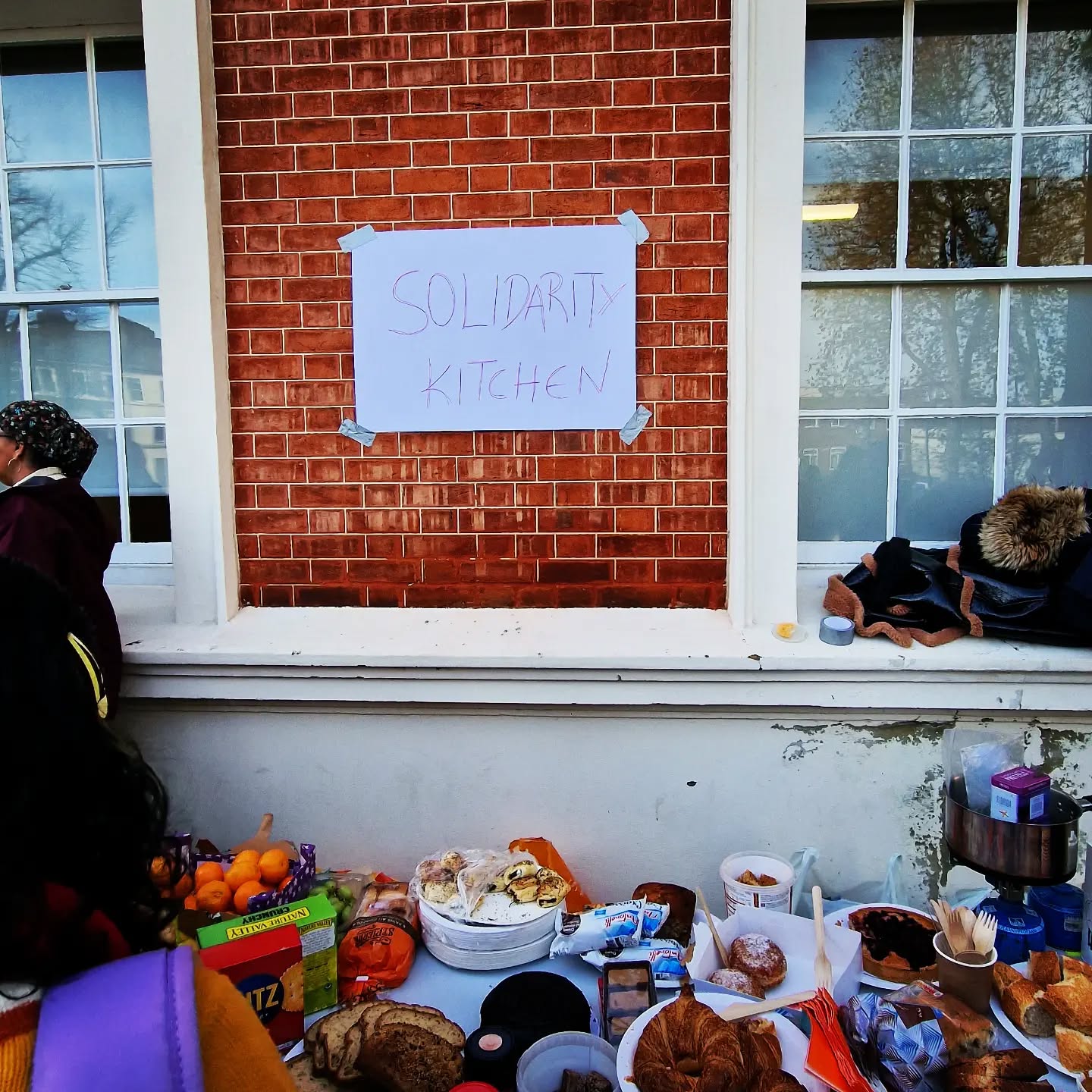 There is a table of assorted food items in the foreground. Behind it is a brick wall with a poster taped to it. The poster says Solidarity Kitchen.