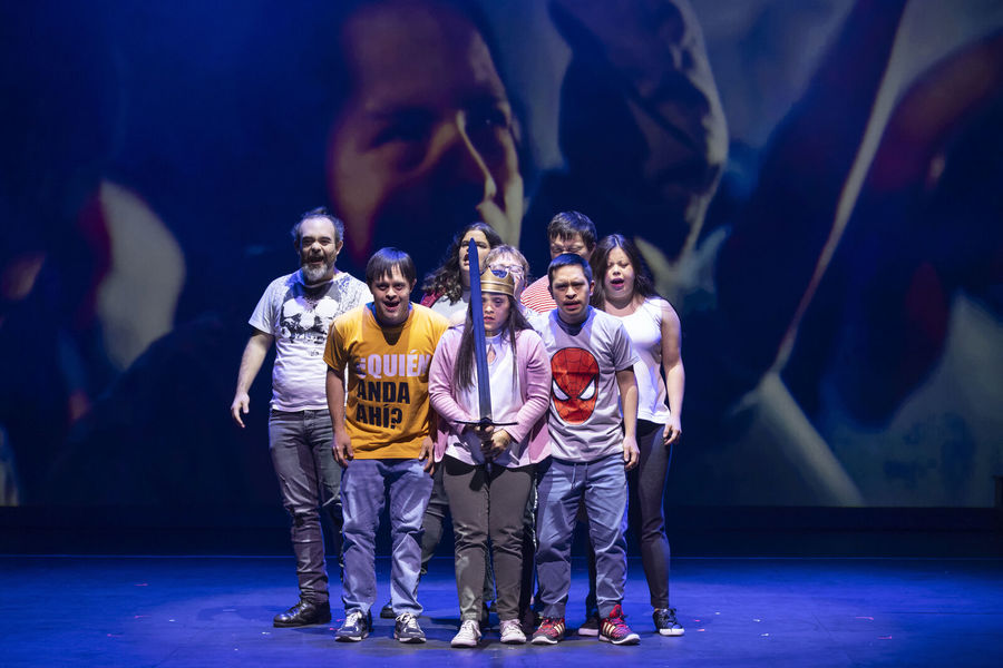 Actors with Down syndrome perform on stage in Hamlet, standing together under blue light as one actress holds a sword at the centre.