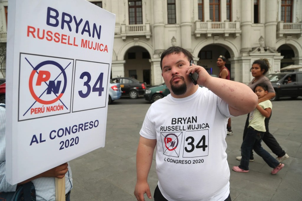 Bryan Russell Mujica, a man with Down syndrome, speaks on the phone in a public square beside a large campaign sign reading “Bryan Russell Mujica, Perú Nación 34, al Congreso 2020.”